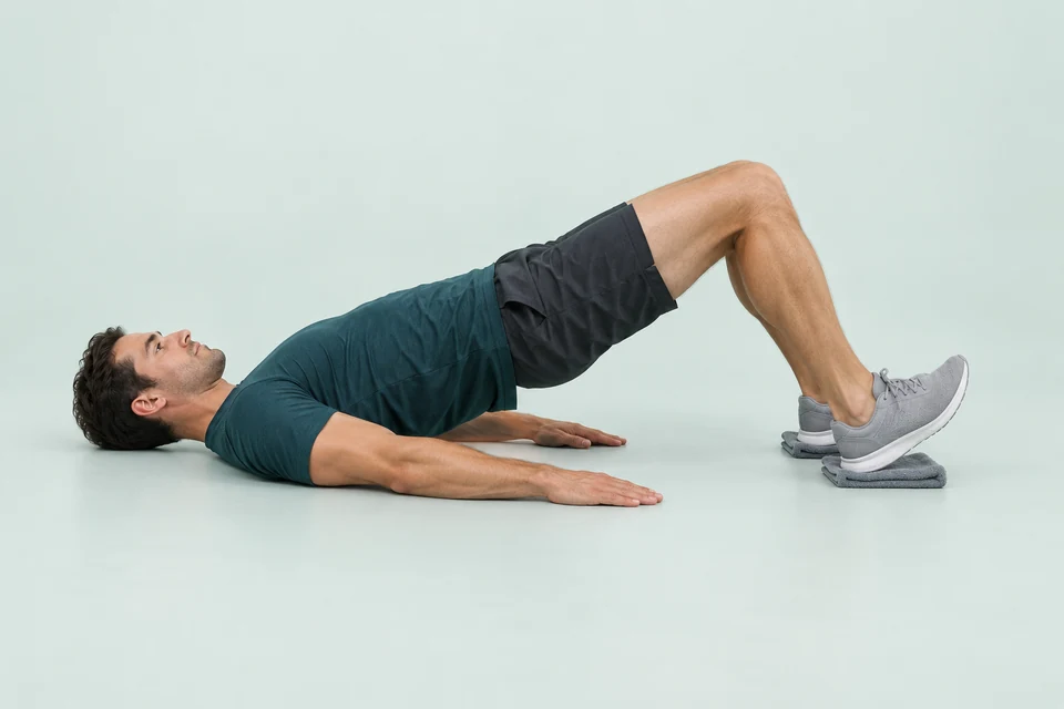 Face-up sliding leg curl with hips raised and both heels resting on separate folded towels.